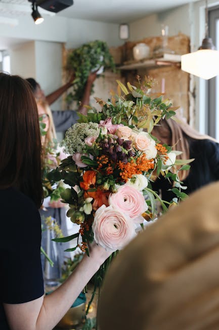 A person holds a large, vibrant floral arrangement featuring soft pink ranunculus, white and pale peach roses, deep purple and orange blooms, and lush green foliage. The bouquet is wrapped with natura