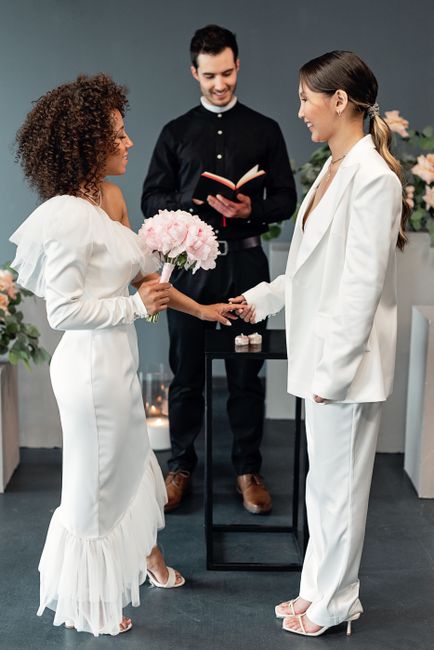 A floral gift exchange taking place indoors, featuring a woman in a white satin dress holding a bouquet of soft pink peonies, and another woman in a white suit warmly shaking her hand. Behind them, a